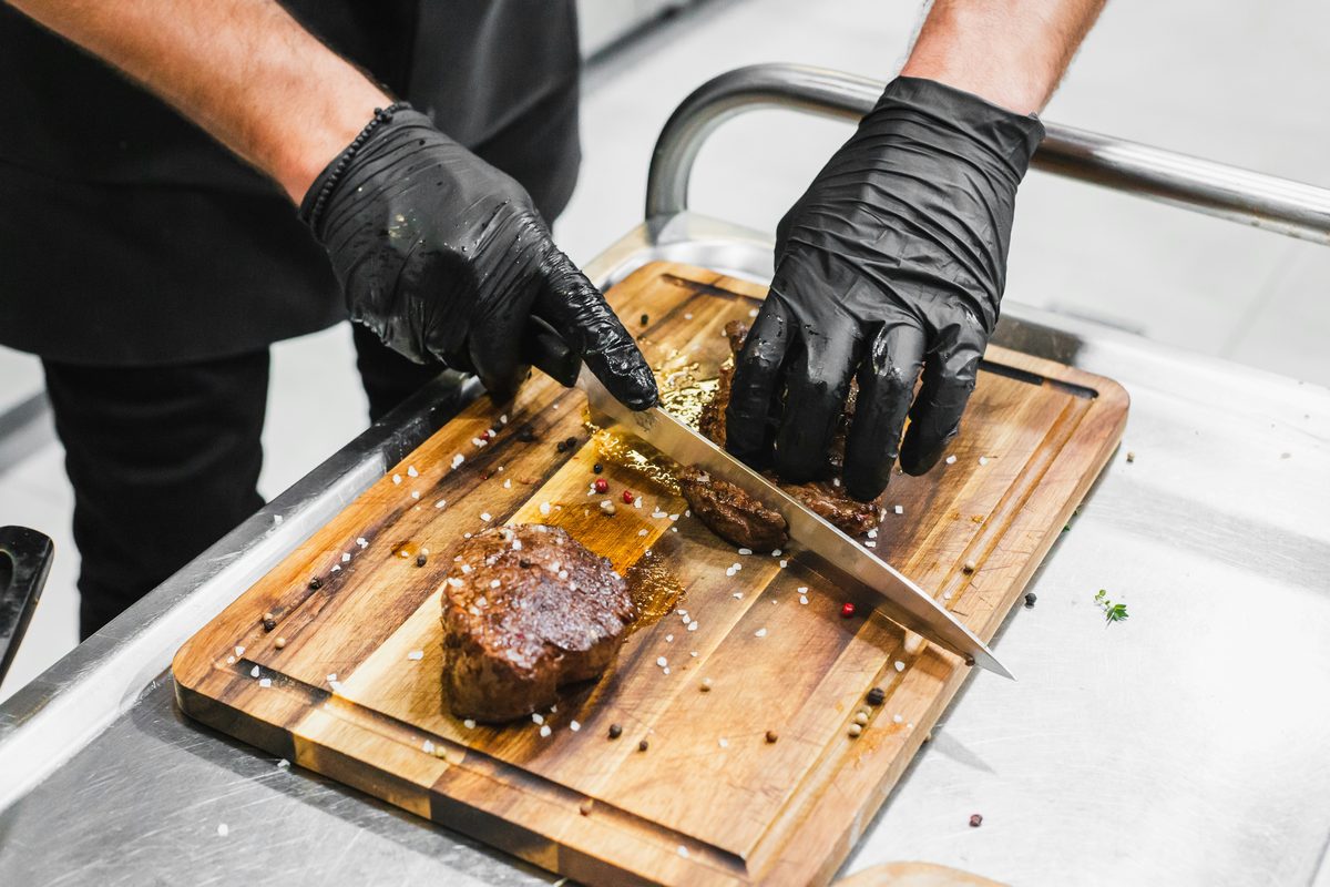 Chef con guantes negros cortando carne cocida sobre tabla de madera — La Carnicerie